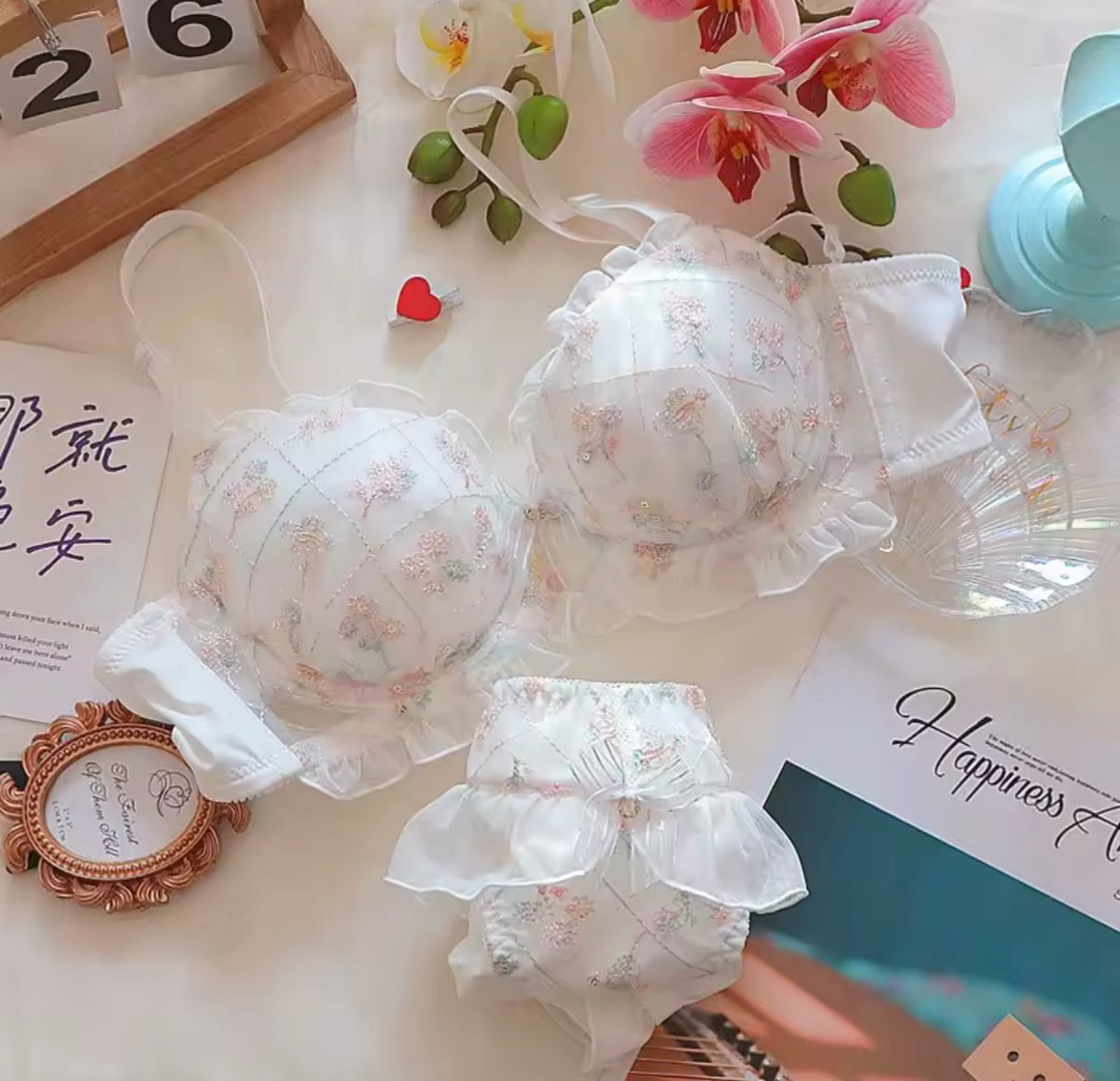 A set of baby lingerie featuring a white ruffled bralette and matching diaper cover with a delicate pastel floral print, styled on a light beige surface with decorative props including pink orchids, a wooden calendar, and stationery.
