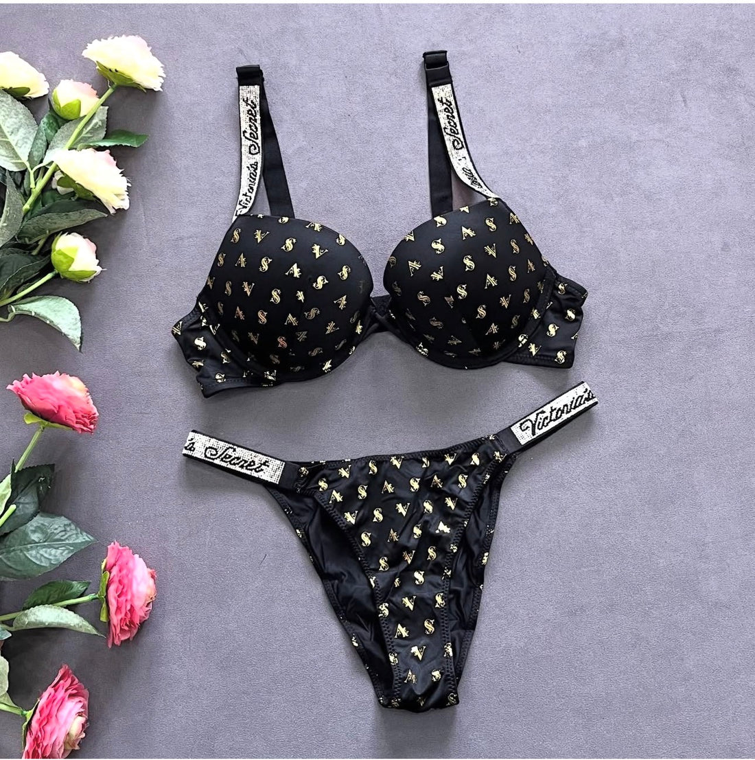 Black lingerie set with gold patterns and 'Victoria's Secret' branding on a gray background with flowers.