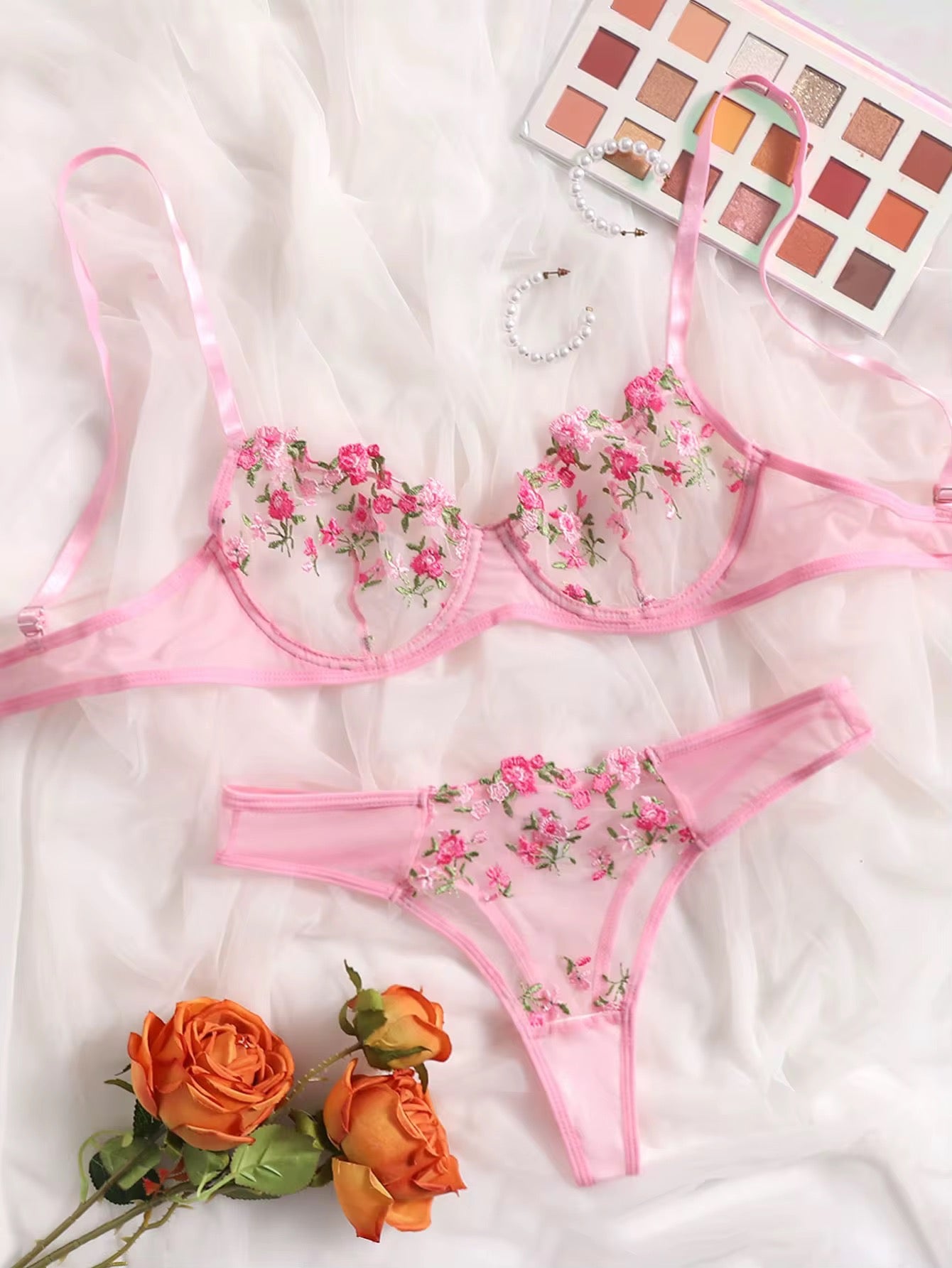 Pink floral lingerie set on a white fabric background with orange roses and a makeup palette.