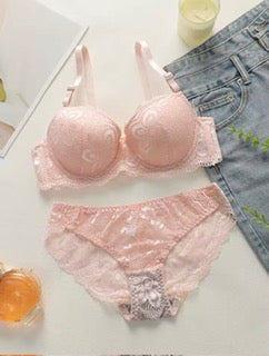 Pink bra and panties set on a white surface with a glass of orange juice and denim jeans in the background.