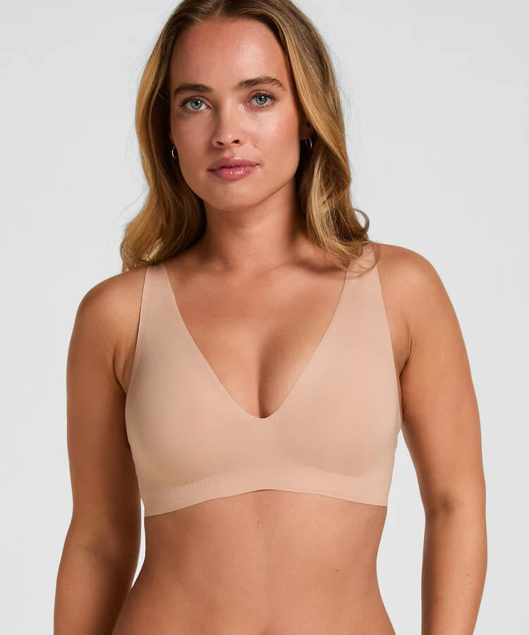 A woman wearing a seamless, scoop-neck bralette in a light beige/nude color, shown from the chest up against a solid light gray background.