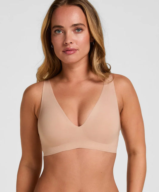 A woman wearing a seamless, scoop-neck bralette in a light beige/nude color, shown from the chest up against a solid light gray background.