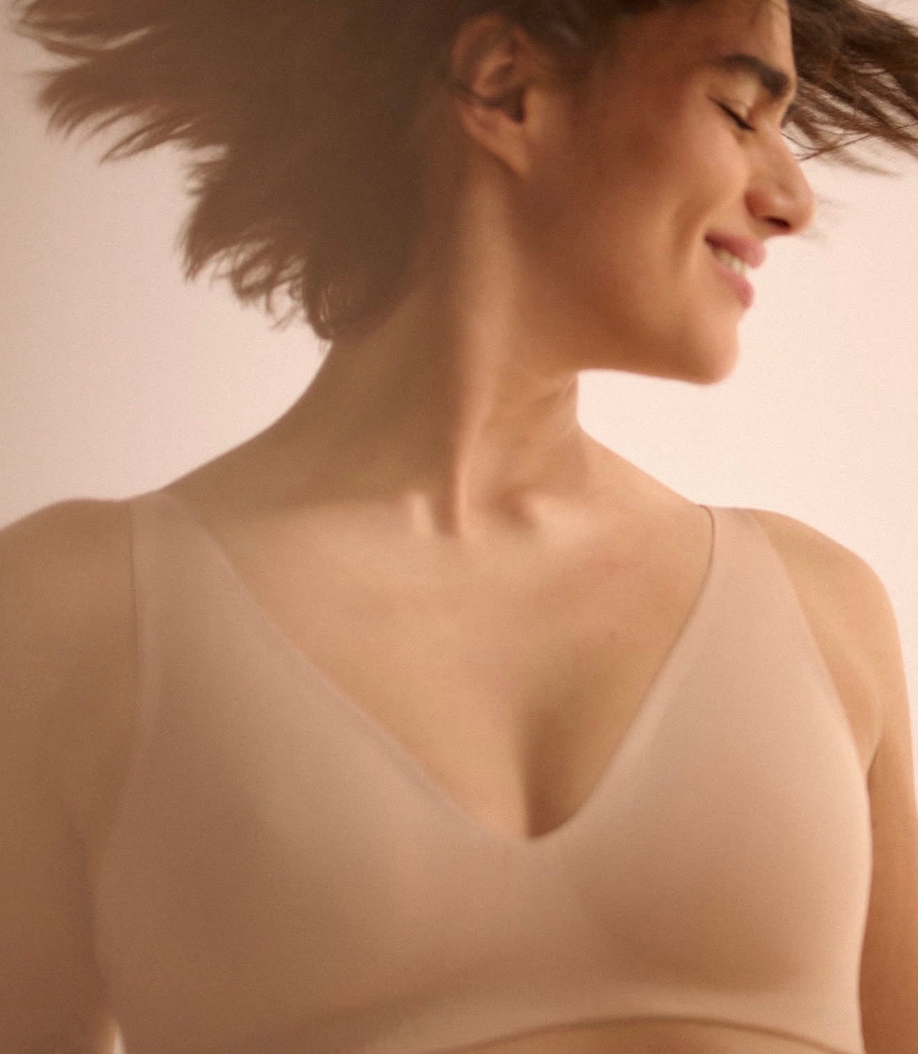 A close-up side profile of a woman wearing a seamless, V-neck bra in a matching nude beige color, with her hair blowing gently in the wind against a soft, light beige background.