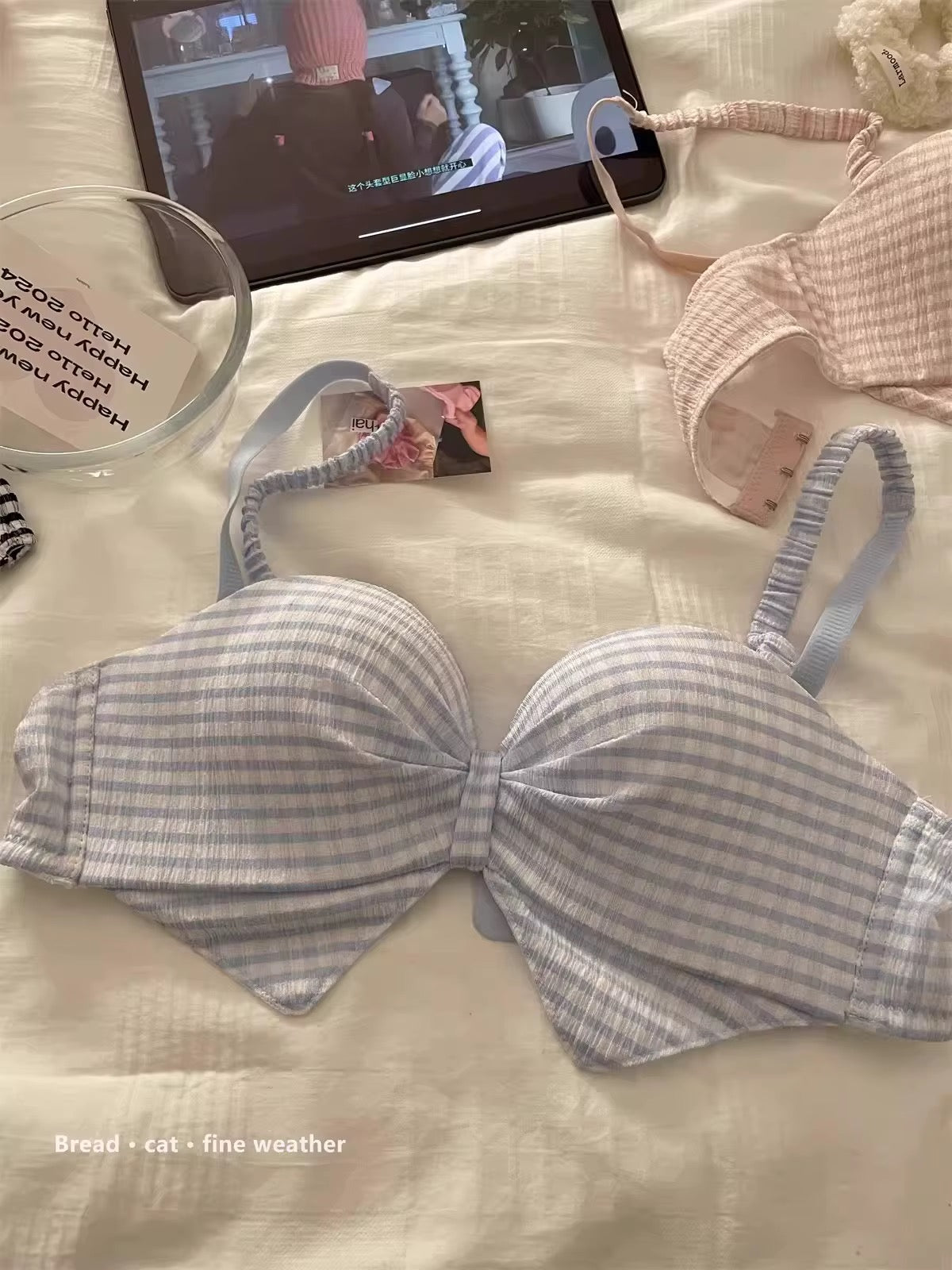 A light blue and white gingham bow-front bra laid flat on a cream-colored fabric surface, with a tablet, a small card, and another pink bra in the background.
