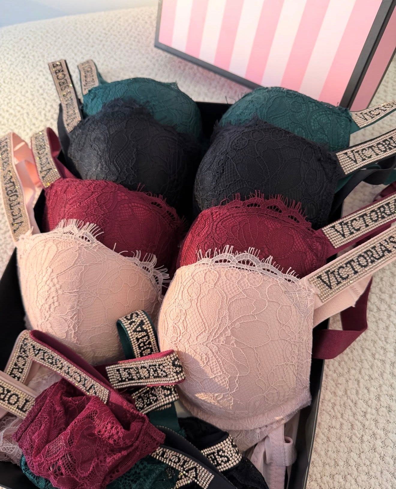 A collection of lace underwire bras in multiple colors, including teal, black, burgundy, and light pink, arranged in a black box with Victoria's brand ribbons. A pink and white striped Victoria's box is partially visible in the background.