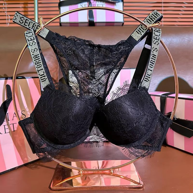 Black lace bra and matching thong set with rhinestone-embellished straps reading 'SECRET', displayed on a gold ring stand with pink-and-white branded boxes in the background.