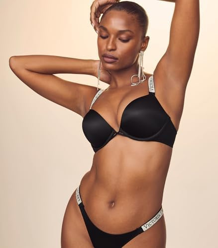 A woman modeling a black bra and matching underwear set with white branded waistband straps against a plain beige studio background.