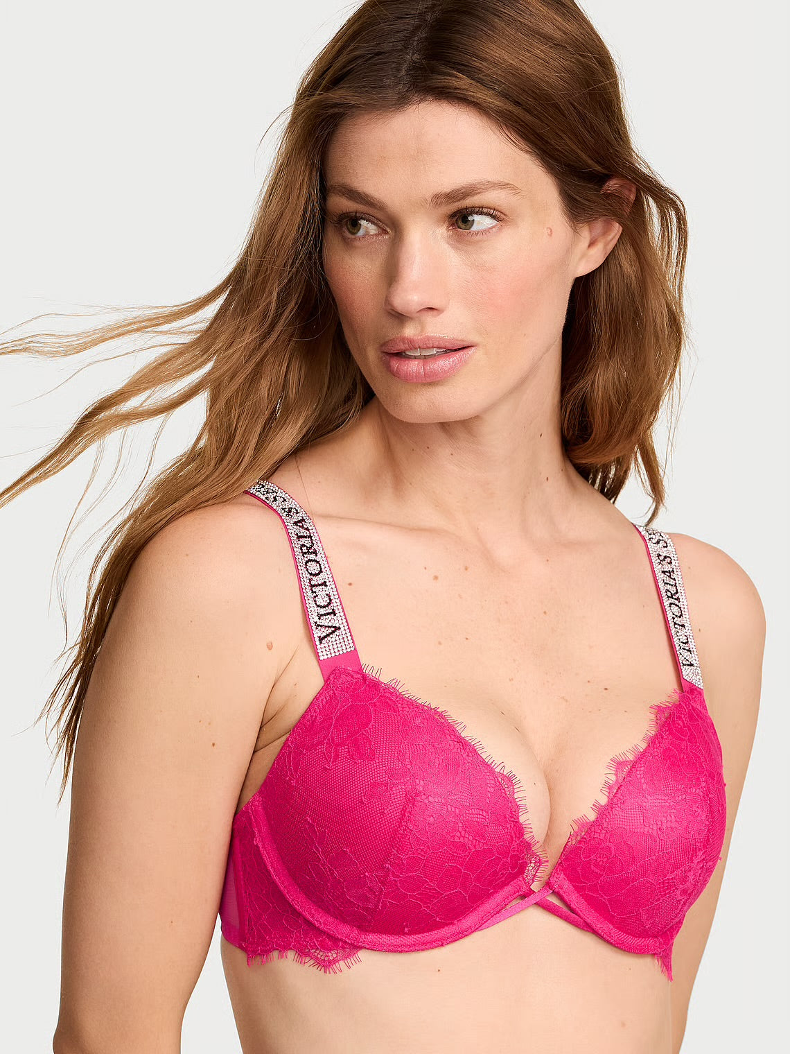 A woman wearing a bright pink lace underwire bra with patterned shoulder straps featuring the brand name Victoria's Secret.