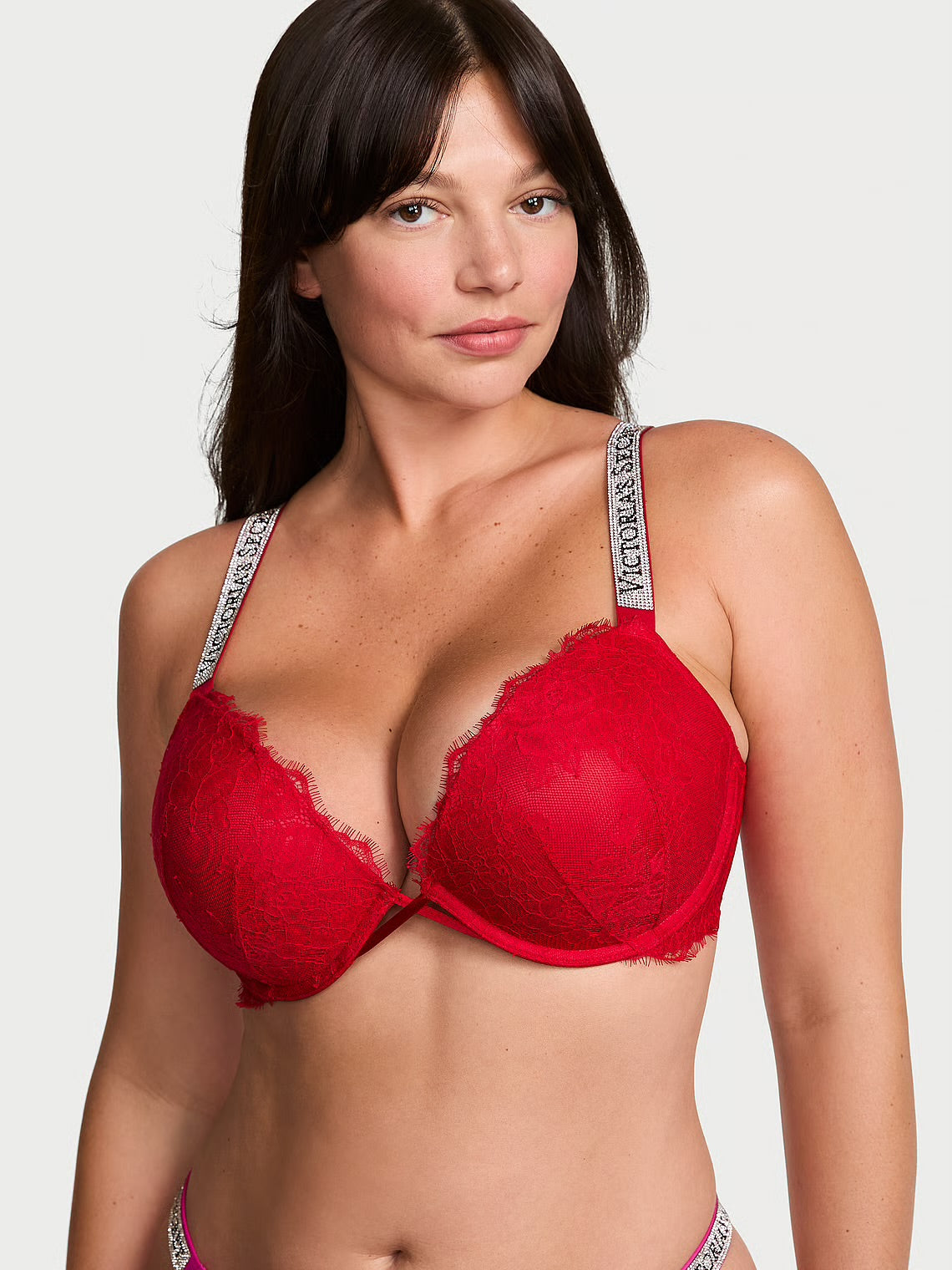 A woman modeling a red lace underwire bra with silver and white Victoria's Secret branded shoulder straps, set against a plain light gray background.