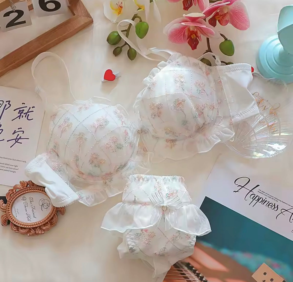 A white ruffled babydoll bra and matching diaper cover set with a delicate pastel floral print, styled in a flat-lay arrangement with decorative props including orchids, a small mirror, and stationery.